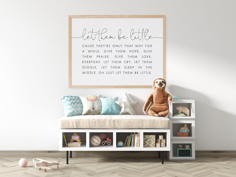 May include: A framed print with a white background and black text that reads "let them be little" and a quote about letting children be children. The print is hanging on a white wall above a white bench with a tan cushion. There are pillows on the bench and a stuffed sloth sitting on the bench.