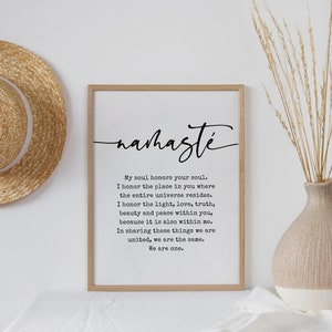 May include: A black and white print with the word "nahaste" in cursive script. The print features a quote about honoring the soul and the universe. The print is framed in a light wood frame and is hanging on a white wall.