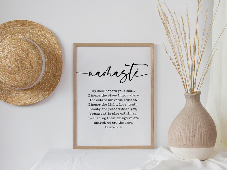 May include: A black and white print with the word "nahaste" in cursive script. The print features a quote about honoring the soul and the universe. The print is framed in a light wood frame and is hanging on a white wall.