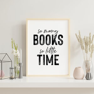 May include: A framed print with the text "So many BOOKS so little TIME" in black brush lettering on a white background. The print is displayed on a white shelf with a vase of white flowers, a candle holder, and a decorative glass vase with dried flowers.