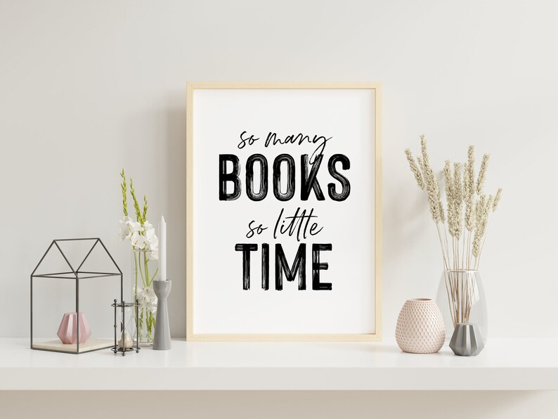 May include: A framed print with the text "So many BOOKS so little TIME" in black brush lettering on a white background. The print is displayed on a white shelf with a vase of white flowers, a candle holder, and a decorative glass vase with dried flowers.
