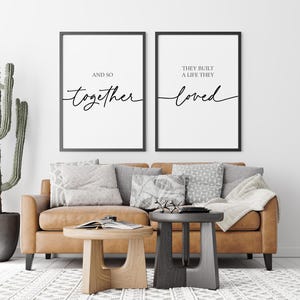 May include: Two black framed prints with white backgrounds and black calligraphy text. The left print reads "And so together". The right print reads "They built a life they loved". The prints are hanging on a white wall above a brown leather sofa with throw pillows.