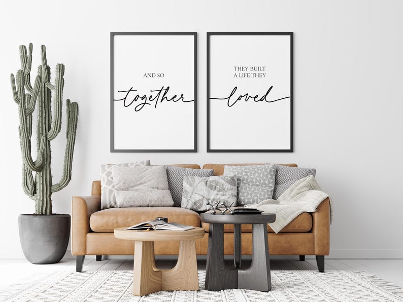 May include: Two black framed prints with white backgrounds and black calligraphy text. The left print reads "And so together". The right print reads "They built a life they loved". The prints are hanging on a white wall above a brown leather sofa with throw pillows.