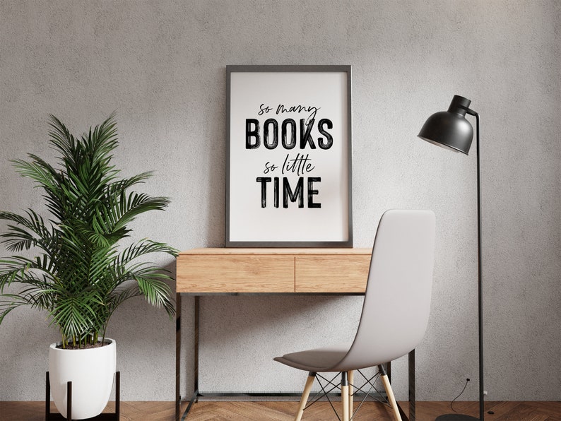 May include: A black framed print with the text "So many books, so little time" hanging on a gray wall above a wooden desk with a white chair and a black floor lamp.