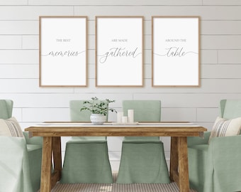 May include: Three framed prints with white backgrounds and black calligraphy text. The prints read "The best memories are made gathered around the table". The prints are hanging on a white shiplap wall above a wooden dining table with four chairs.