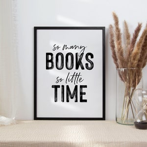 May include: A black framed print with the text "So many BOOKS so little TIME" in white lettering. The print is sitting on a beige table with a vase of dried pampas grass and a woven basket.