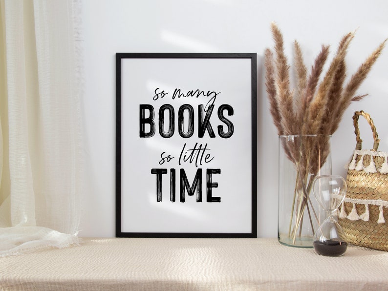 May include: A black framed print with the text "So many BOOKS so little TIME" in white lettering. The print is sitting on a beige table with a vase of dried pampas grass and a woven basket.