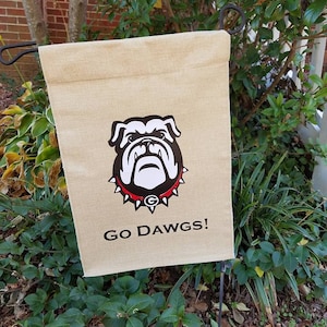 May include: A beige burlap garden flag with a black and white bulldog illustration. The bulldog has a red and black spiked collar with a white "G" in the center. The text "GO DAWGS!" is printed in black below the bulldog.