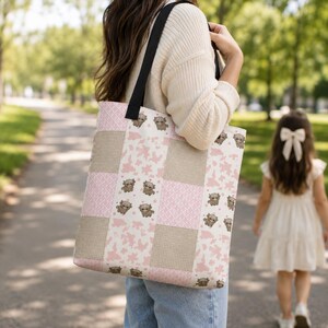 May include: A patchwork tote bag with a black strap. The bag features a pattern of pink cow spots, brown squares, and cartoon Highland cows. The bag is carried by a person walking on a path.