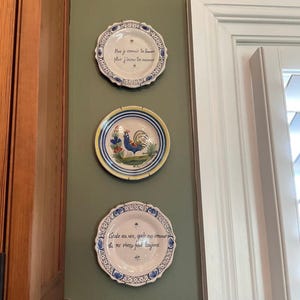 May include: Three decorative ceramic plates mounted vertically. The top plate features blue floral borders and French text. The middle plate displays a colorful rooster illustration with blue and yellow stripes. The bottom plate has blue geometric patterns and French text.