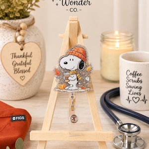 May include: A glittery acrylic badge reel featuring a cartoon dog wearing an orange beanie and a yellow scarf, holding a coffee cup. The character is surrounded by autumn leaves and a small pumpkin. The badge reel is displayed on a wooden easel.