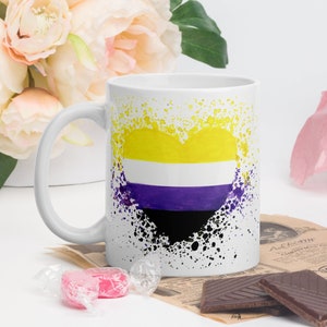 Puede incluir: Taza de cerámica blanca con un diseño de corazón negro, morado, blanco y amarillo. El corazón está rodeado de salpicaduras de pintura negra.