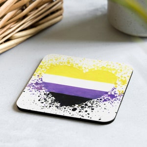 Puede incluir: Un posavasos cuadrado con fondo blanco y un diseño en forma de corazón negro, morado, blanco y amarillo con efecto de pintura salpicada.
