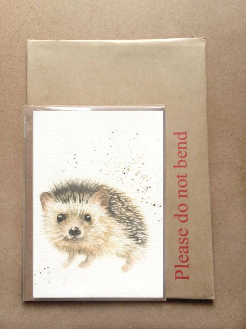 May include: A watercolor painting of a hedgehog on a white card. The hedgehog is brown and gray with a white belly. The card is surrounded by a brown envelope with the words "Please do not bend" printed on it.