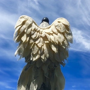 Puede incluir: Un disfraz de alas de &aacute;ngel de fieltro blanco con ribete de encaje, exhibido en un maniqu&iacute; sobre un fondo de cielo azul.