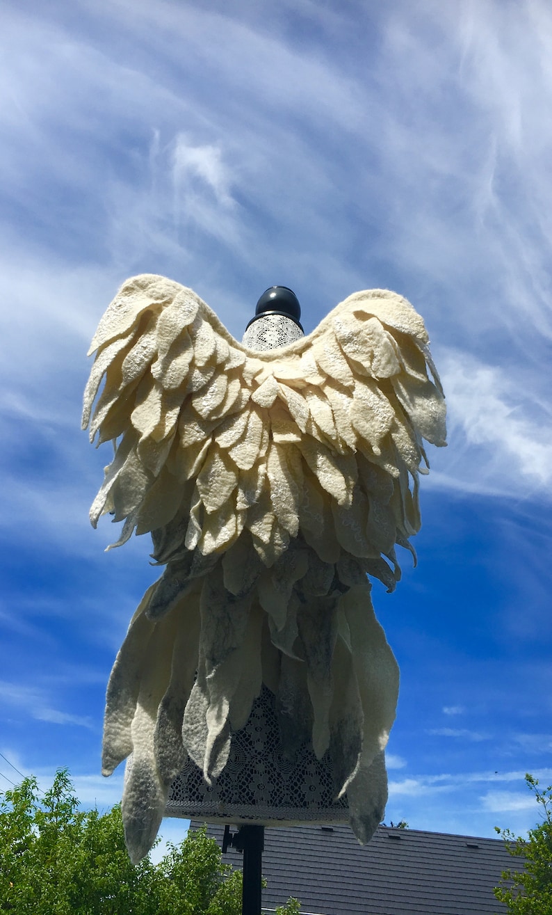 Puede incluir: Un disfraz de alas de &aacute;ngel de fieltro blanco con ribete de encaje, exhibido en un maniqu&iacute; sobre un fondo de cielo azul.