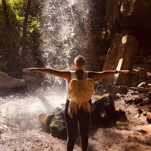 Puede incluir: Una persona con una camisa blanca y pantalones negros con grandes alas de &aacute;ngel de plumas se encuentra frente a una cascada.