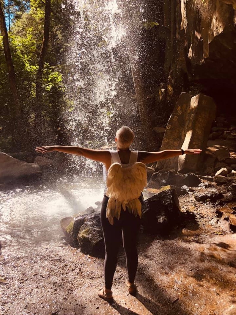 Puede incluir: Una persona con una camisa blanca y pantalones negros con grandes alas de &aacute;ngel de plumas se encuentra frente a una cascada.