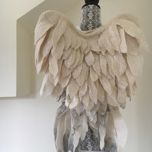 Puede incluir: Un par de alas de &aacute;ngel de fieltro blanco con un efecto degradado gris en la parte inferior. Las alas est&aacute;n unidas a un maniqu&iacute; cubierto de encaje blanco.