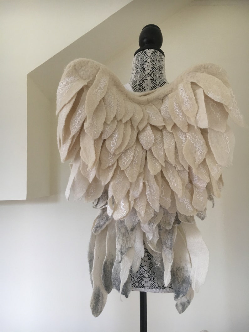 Puede incluir: Un par de alas de &aacute;ngel de fieltro blanco con un efecto degradado gris en la parte inferior. Las alas est&aacute;n unidas a un maniqu&iacute; cubierto de encaje blanco.