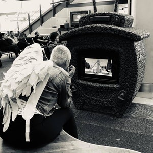 Puede incluir: Una persona con un disfraz de alas de &aacute;ngel blancas se sienta frente a una gran escultura en forma de televisor, negra y texturizada. La escultura tiene una pantalla que muestra una imagen de dibujos animados de una casa con un campanario de iglesia.