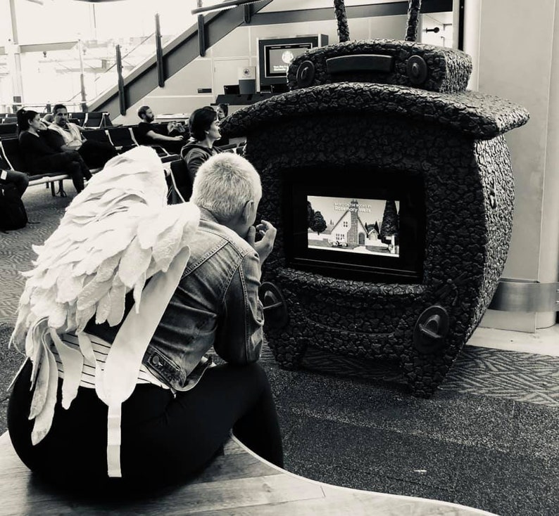 Puede incluir: Una persona con un disfraz de alas de &aacute;ngel blancas se sienta frente a una gran escultura en forma de televisor, negra y texturizada. La escultura tiene una pantalla que muestra una imagen de dibujos animados de una casa con un campanario de iglesia.