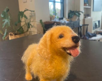 May include: A needle felted golden retriever dog figurine. The dog is standing on a dark grey surface and has a pink tongue sticking out.