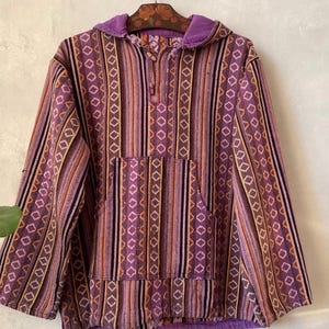 May include: A long-sleeve hooded pullover featuring a vertical striped pattern in shades of purple, orange, and black. The garment includes a front kangaroo pocket, a drawstring neckline, and a solid purple hood lining.