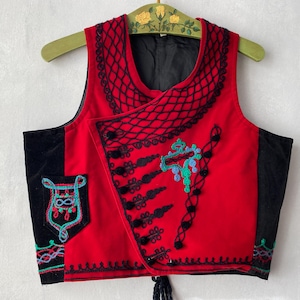 May include: A sleeveless red waistcoat featuring black trim, a black mesh-like pattern around the neckline, and black decorative buttons. The garment includes black side panels, a small pocket with teal and pink embroidery, and additional black cord detailing.