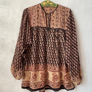 May include: A long-sleeved blouse featuring a dark brown and tan paisley print. The garment has a round neckline with a central slit and tie closure, a yoke panel with vertical patterns, and a wide horizontal border at the hemline.