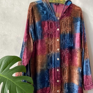 May include: A long-sleeved hooded shirt made of crushed velvet in a patchwork pattern of blue, magenta, and copper tones. The front features a vertical button-down closure with white buttons and decorative embroidery flanking the placket.