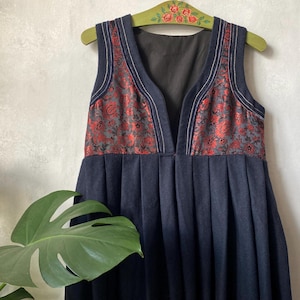 May include: A sleeveless navy blue dress featuring a deep V-neckline and a pleated skirt. The bodice is accented with panels of red and black floral brocade fabric. The dress is displayed on a green wooden hanger decorated with painted red roses.