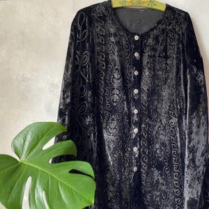May include: A black velvet long-sleeve button-down shirt featuring vertical panels of intricate tonal embroidery. The garment has a round neckline and a front placket with small round buttons. The fabric has a crushed texture throughout.
