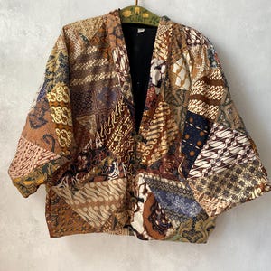May include: A patchwork jacket featuring a variety of brown, tan, and navy blue batik patterns. The garment has a V-neckline, wide sleeves, and a front closure with three circular buttons. The interior of the jacket is lined with solid black fabric.