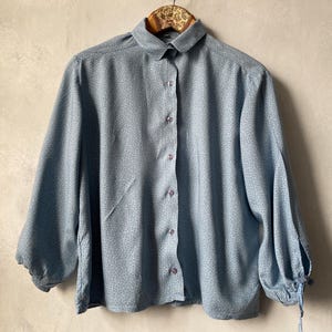 Könnte beinhalten: Ein hellblaues Langarmhemd mit einem kleinen, sich wiederholenden dunkelblauen Blumenmuster. Das Hemd hat einen abgerundeten Kragen, eine Knopfleiste auf der Vorderseite und Ärmel, die in Manschetten mit kleinen Bindebändern enden.