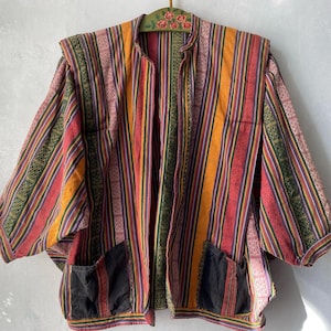 Könnte beinhalten: Eine Jacke mit einem lebendigen vertikalen Streifenmuster in Orange-, Rot-, Grün- und Violetttönen. Das Kleidungsstück hat weite Ärmel, einen Stehkragen und zwei dunkel gefärbte aufgesetzte Taschen auf der Vorderseite. Der Stoff zeigt durchgehend komplexe, gewebte geometrische Texturen.