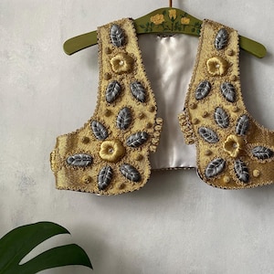 Puede incluir: Un chaleco sin mangas con una base de tela dorada metálica y bordados intrincados. El diseño incluye motivos florales dorados en relieve y adornos en forma de hoja de color gris plateado. El interior está forrado con satén blanco roto.