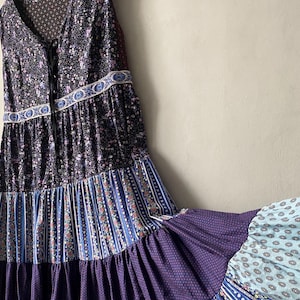 Pode incluir: Um vestido sem mangas com camadas, apresentando um corpete preto com estampa floral pequena e cordões na frente. A saia consiste em vários painéis horizontais com padrões variados, incluindo listras azuis e brancas, flores e pontos roxos.