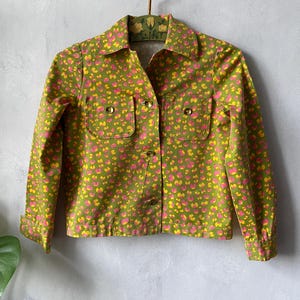 May include: A long-sleeve button-up jacket in an olive green base color featuring a repeating pattern of small, irregular pink and yellow spots. The jacket includes a pointed collar, two chest pockets with button closures, and buttoned cuffs.