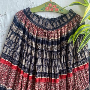 May include: A patterned pleated skirt in navy blue, red, and beige. The skirt has a repeating floral and paisley design. The skirt is hanging on a green wooden hanger with floral detailing.