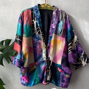 May include: A multi-coloured patchwork jacket featuring a vibrant array of purple, blue, pink, and teal panels with abstract patterns. The jacket has three dark buttons down the front, wide three-quarter length sleeves, and a solid black interior lining.