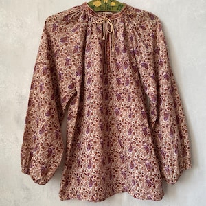 Könnte beinhalten: Eine langärmelige Bluse mit einem dichten Blumen- und Paisleymuster in Braun-, Beige- und gedämpften Violetttönen. Das Kleidungsstück hat einen Rundhalsausschnitt mit zentralem Bindeband sowie geraffte Stoffdetails an Schultern und Ärmelbündchen.