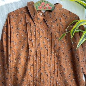 Può includere: Una camicia a maniche lunghe marrone con un motivo ripetuto di forme paisley blu scuro e piccoli accenti floreali arancioni. Il capo presenta un colletto a punta e un pannello frontale dettagliato con pieghe verticali centrali.
