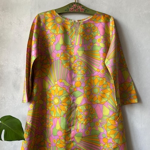 May include: A long-sleeved dress featuring a vibrant geometric pattern in shades of lime green, bright pink, and golden yellow. The garment has a round neckline and is displayed on a green wooden hanger decorated with small painted pink roses.