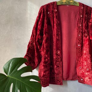 Peut inclure: Une veste en velours rouge profond avec des manches longues et une ouverture sur le devant. Le vêtement est orné de broderies ton sur ton complexes et de petits miroirs circulaires sur les panneaux avant et les manches, créant un aspect texturé.