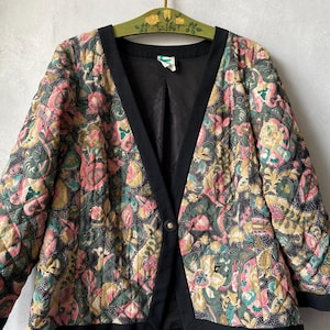 Könnte beinhalten: Eine gesteppte Jacke mit einem mehrfarbigen Blumen- und Paisle Blumen- und Paisley-Muster in Rosa-, Grün- und Gelbtönen auf dunklem Grund. Die Jacke hat einen einfarbigen schwarzen Besatz entlang des V-Ausschnitts, der Ärmelbündchen und des Saums mit einem Knopf.