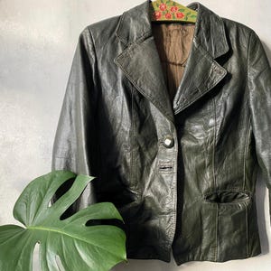 Puede incluir: Una chaqueta tipo blazer de cuero en color verde oliva oscuro con solapa de muesca, cierre frontal de un solo botón y dos bolsillos laterales. La prenda tiene un forro interior marrón y cuelga de una percha de madera con flores pintadas.