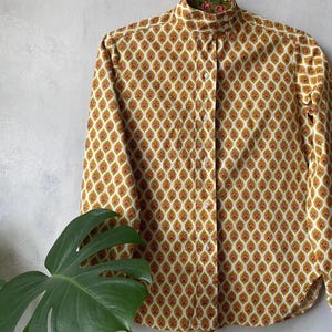 May include: A long-sleeved button-down shirt with a high collar. The shirt features a repeating pattern of gold and red teardrop shapes on a cream background. The collar lining has a floral pattern.
