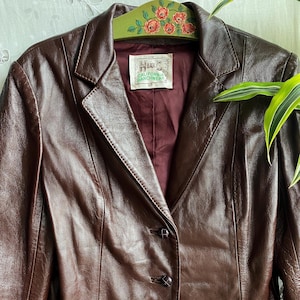 Può includere: Un blazer in pelle marrone scuro con revers dentellato, due bottoni frontali e una fodera color bordeaux intenso. Un'etichetta interna bianca riporta la scritta H&C CALIFORNIA RANCHWEAR. Il capo è appeso a una gruccia verde con motivo floreale.