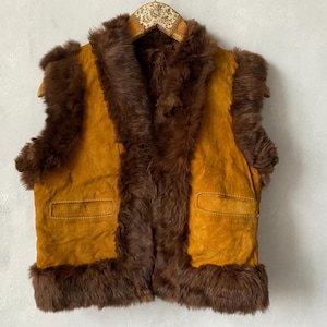 Puede incluir: Un chaleco vintage de ante en amarillo mostaza cálido con ribete de piel marrón. El chaleco tiene dos bolsillos delanteros y un cuello de piel marrón. El chaleco está colgado de una percha de madera.